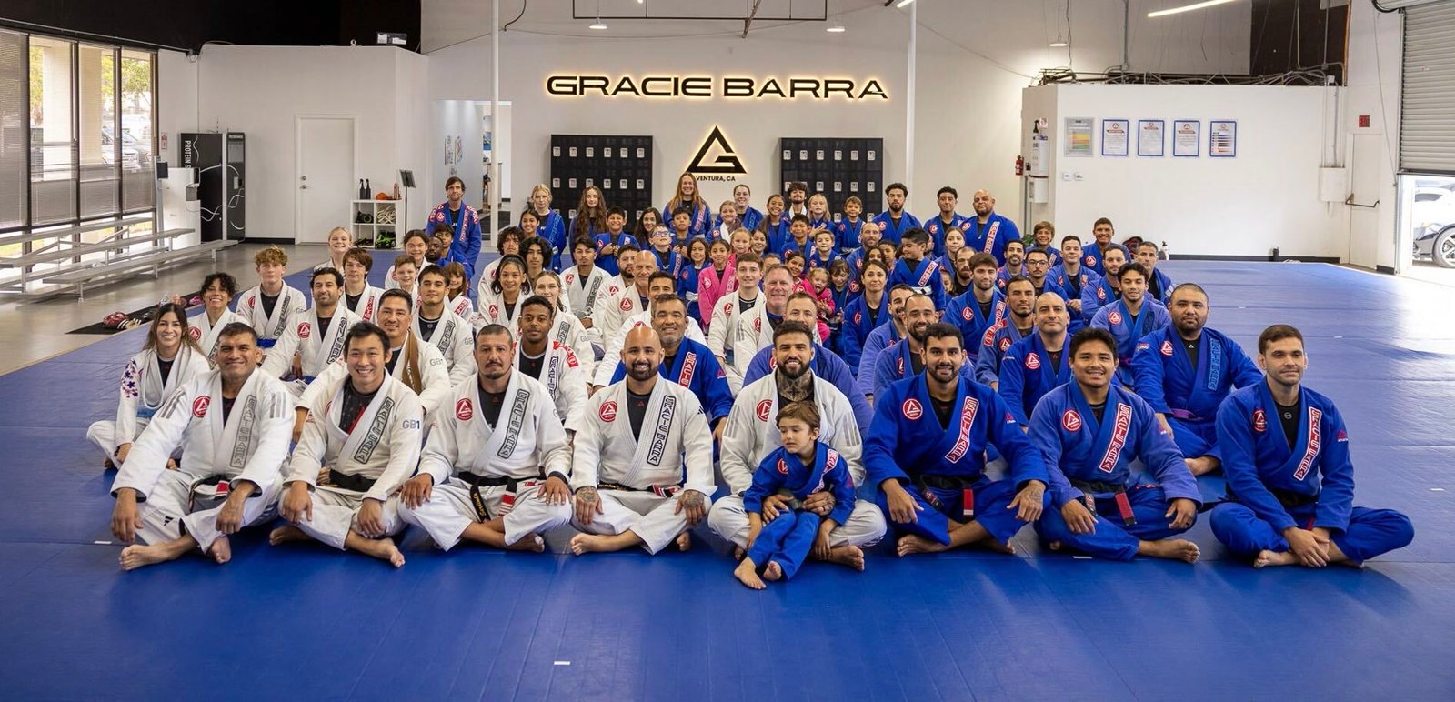 The full Gracie Barra Ventura academy, kids and adults in white and blue gis, seated together on the blue mats